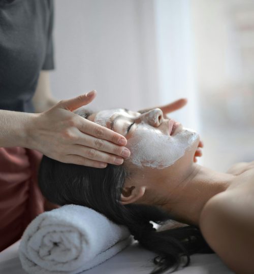 Faceless beauty master smearing mask on clients face while woman relaxing on massage table and getting spa procedure